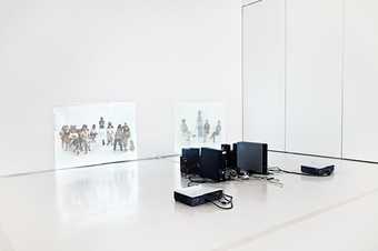 A video installation: two projections of groups of people on white walls with the projecting equipment in the centre of the room