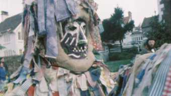 video still of person wearing a mask and a large costume made of strips of fabric
