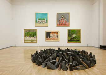 An interior of an art gallery. A circle of black stones on the wooden floor. Five paintings of landscape and architectural forms on the wall behind.