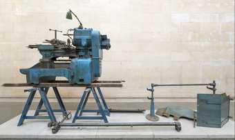 A piece of heavy machinery on a trestle bench on a plinth in a gallery