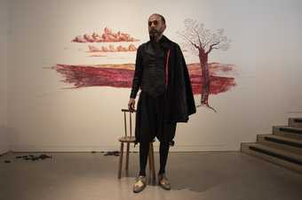 Artist Nikhil Chopra stands in front of his mural, drawn in different shades of red lipstick on a white wall. Chopra wears heavy eyeliner, and is dressed with reference to an Elizabethan bard in a corseted jacket, velvet cloak, tights, and heeled shoes.