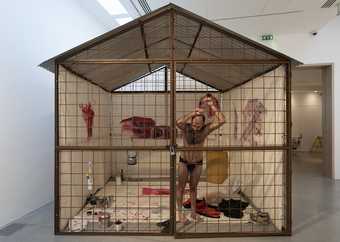 Nikhil Chopra, wearing only black underpants, leans against a wire cage that covers one side of a single-room structure, constructed inside a gallery space. The interior walls are covered in red murals; paint pots and clothing are scattered on the floor.