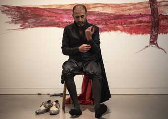 Nikhil Chopra sits on a chair in front of his red mural, his shoes removed and set to one side, one arm raised as he unbuttons one cuff. His gaze is directed to the floor, appearing contemplative.