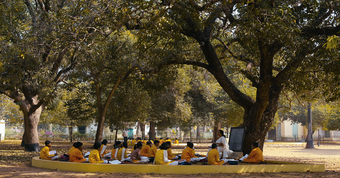 A class of students wearing bright yellow uniform sit in front of a teacher under a tree