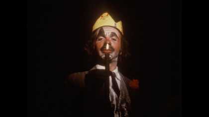 a man with clown-like make up on stands with a crown