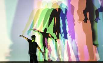 A person standing facing a wall with arms outstretched. On the wall their shadow is cast in multiple different colours.