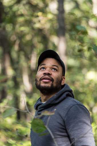 Portrait of the artist Oscar Murillo wearing a hat and looking up to the sky. behind him are trees