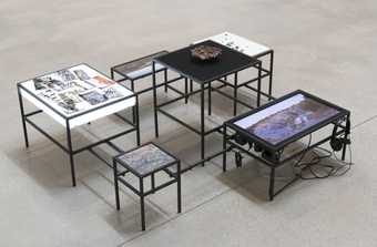 Installation shot of a few low table-like forms with video, photography and found materials on them