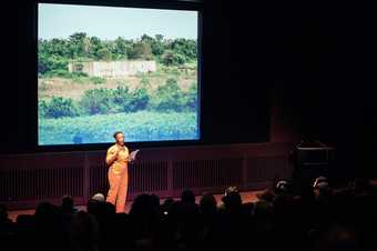 Artist Otobong Nkanga stands onstage in the Starr Cinema at Tate Modern, wearing an orange jumpsuit, reading from a piece of paper and speaking to the audience. Behind her is a projected image of a Nigerian landscape with the ruins of a concrete structure