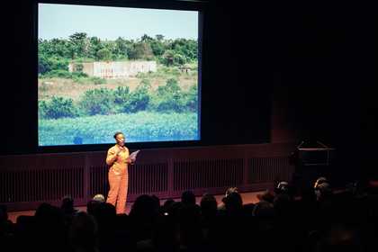 Artist Otobong Nkanga stands onstage in the Starr Cinema at Tate Modern, wearing an orange jumpsuit, reading from a piece of paper and speaking to the audience. Behind her is a projected image of a Nigerian landscape with the ruins of a concrete structure