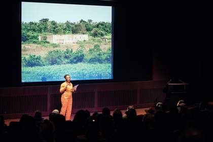 Artist Otobong Nkanga stands onstage in the Starr Cinema at Tate Modern, wearing an orange jumpsuit, reading from a piece of paper and speaking to the audience. Behind her is a projected image of a Nigerian landscape with the ruins of a concrete structure
