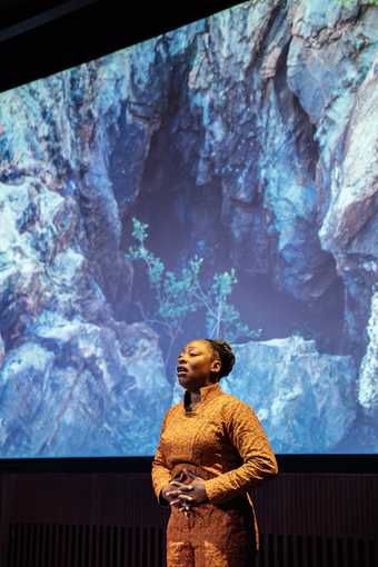 Artist Otobong Nkanga stands with her hands on her abdomen, fingers interlaced, breathing deeply with chin raised. Behind her is a projected image of a rocky landscape