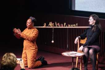 Photograph of two people on stage; one is artist Otobong Nkanga, who kneels with arms outstretched towards the audience, fingertips almost touching. Behind Nkanga, Anne Barlow sits on a chair and watches.