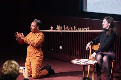 Photograph of two people on stage; one is artist Otobong Nkanga, who kneels with arms outstretched towards the audience, fingertips almost touching. Behind Nkanga, Anne Barlow sits on a chair and watches.