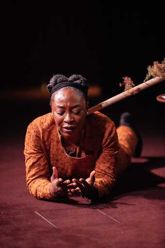 Otobong Nkanga lies on the stage, on her front, resting on elbows, palms upwards. She looks down at her hands, her mouth open as if speaking, a pained or worried look on her face. The horizontal pole with model trees is visible to the right of her head