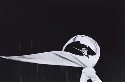 black background with a figure dressed in white in the foreground. One arm is curved over their head and the second poitning away from their body.