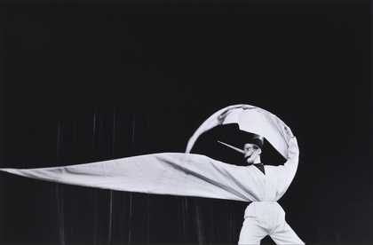 black background with a figure dressed in white in the foreground. One arm is curved over their head and the second poitning away from their body.