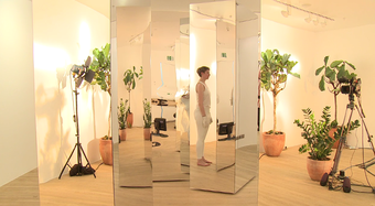 Image shows a room full of mirrors and plants and a woman standing in white leggings side on
