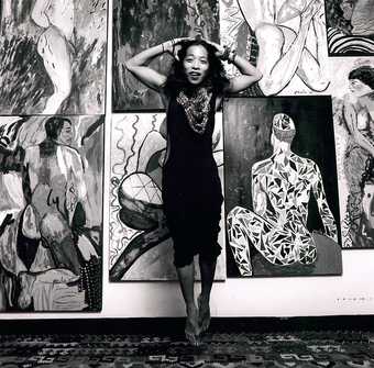 monochrome photo of artist Pacita Abad posing in front of a wall of paintings