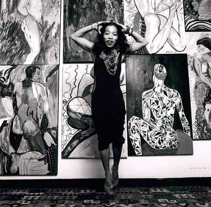 monochrome photo of artist Pacita Abad posing in front of a wall of paintings