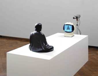 statue of buddha sat on plinth facing a small tv screen and camera