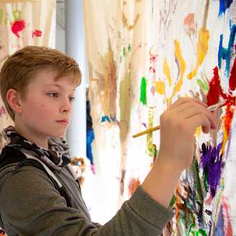 A young boy is standing as he paints some hanging material with colourful paints