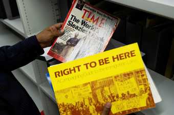 Hands hold up two magazines/pamphlets by Library shelves: one is Time magazine with headline 'The World's Sleaziest Bank'; the other is yellow with the title 'Right To Be Here: A Campaigning Guide to the Immigration Laws'.