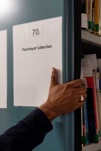 A hand reaches around book stacks in the Tate Library; the shelf is labelled '70. Panchayat Collection'