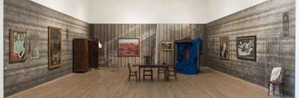 Installation view of an empty stage which is set up like a living or dining room with lots of artworks hanging on the walls
