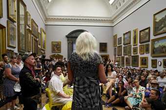 A speaker with a microphone addresses a group of people gathered sitting on the floor in the Tate Britain 1840s room.