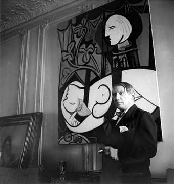 Black and white photograph of Picasso stood beside a painting