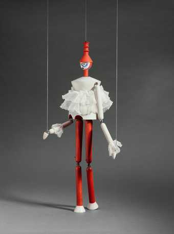 A wooden marionette puppet held up by two strings wears a red hat, a white ruffled top and red trousers 