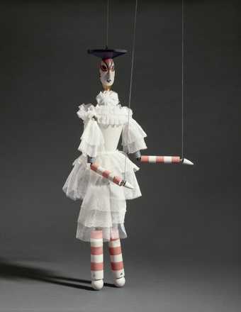 A marionette with stripey arms and legs and a white ruffled dress 