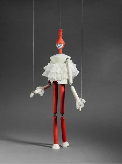 A wooden marionette puppet held up by two strings wears a red hat, a white ruffled top and red trousers