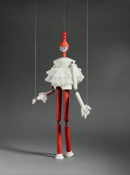 A wooden marionette puppet held up by two strings wears a red hat, a white ruffled top and red trousers 