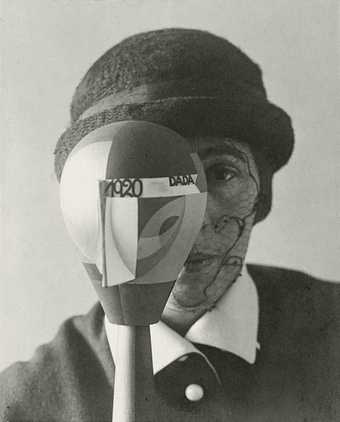 A black and white photograph where a woman faces the camera with a wooded abstract head sculpture in front of one side of her face. The woman wears a bowler hat and has face paint on the visible side of her face.