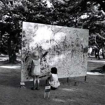 Please Draw Freely, situated outside in a woods a large wooden board is drawn on by two small children