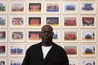 landscape photograph showing the artist Steve McQueen in front of his Year 3 project. Behind him are many framed photographs of school groups. He is wearing glasses and a black jacket.