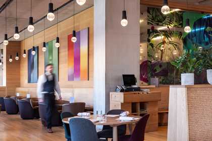 a waiter is walking through a restaurant . there are a row of tables and chairs with bright colourful artworks on the wall behind. from the ceiling bare light bulbs hang down..