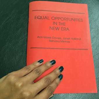 Photograph of a hand with painted nails resting on the cover of a red pamphlet titled Equal Opportunities in the New Era