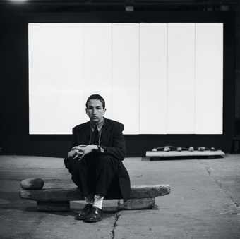 Photograph of Robert Rauschenberg seated on Untitled (Elemental Sculpture) with White Painting (seven panel) behind him at the basement of Stable Gallery, New York 1953