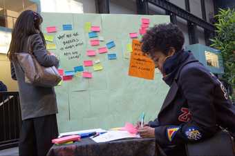 Two people contribute ideas to a board of colourful sticky notes, responding to the question 'What are the barriers to working in arts and culture?'