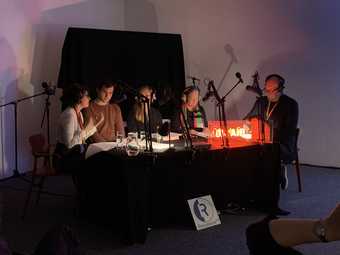 Five people sit around a table covered in microphones on stands, the room is dark and a red light glows on the table