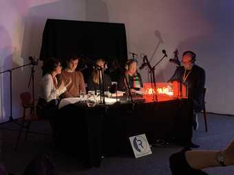 Five people sit around a table with many microphones on stands, the room is dark except for a glowing red light on the table. A sign reads 'Resonance FM'