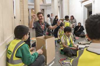 Photograph of a schools workshop at Tate Britain. Young students wearing hi-vis jackets making wooden sculptures in the gallery 