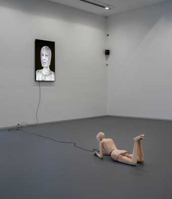 installation view of a wooden figure lying down in a gallery space with digital screens