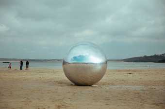 A large silver ball stands alone next to a grey sea under a stormy grey sky
