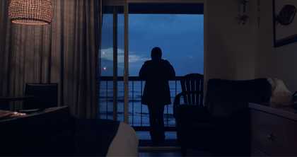 A woman stands on a balcony overlooking the sea at sunset