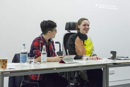 Two speakers sit at a table with a projected screen behind them