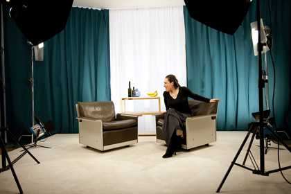 A performer sits on an armchair in the set of a shoot lit with spotlights